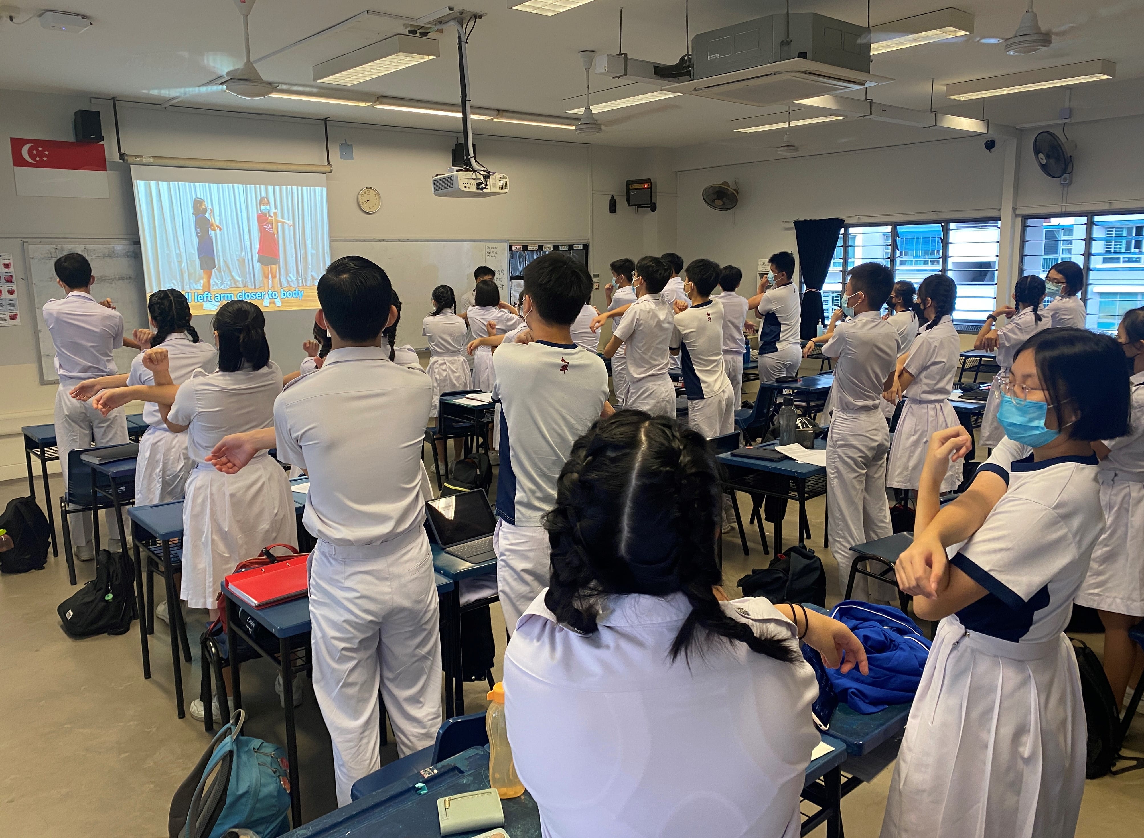 Students in white uniforms stand at desks following exercise movements shown on a projected video screen in a classroom.